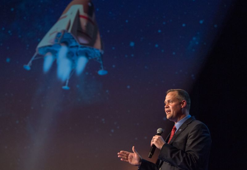 A man in a suit speaks into a microphone in front of a large image of a spacecraft.