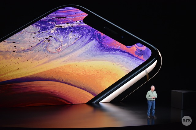 A man on a stage is dwarfed by a gigantic video display of a smartphone.