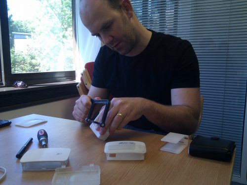 Phone photo of Eben sitting at a desk and hacksawing a white 3D-printed prototype Raspberry Pi case