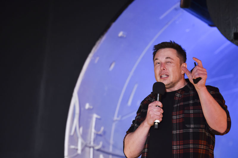 A casually dressed man gestures while speaking into a microphone.