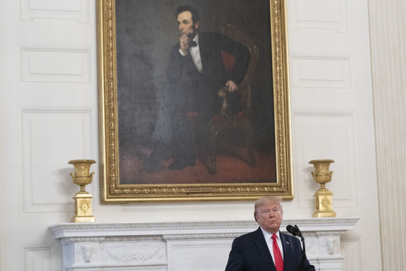 A man speaks from a podium while being dwarfed by a painting of Abraham Lincoln.