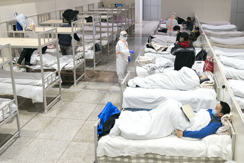 A tech in protective gear inspects patients in a row of beds.