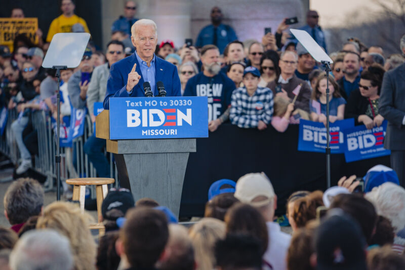 A man in an open-collared suit speaks into a microphone.