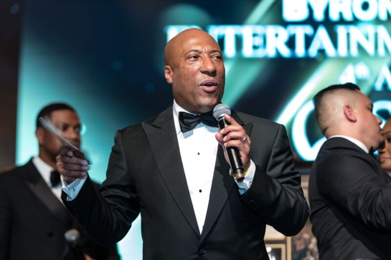 Byron Allen wearing a tuxedo and holding a microphone while speaking on stage at a charity event.