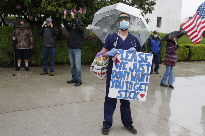A masked man in OR scrubs holds a sign saying