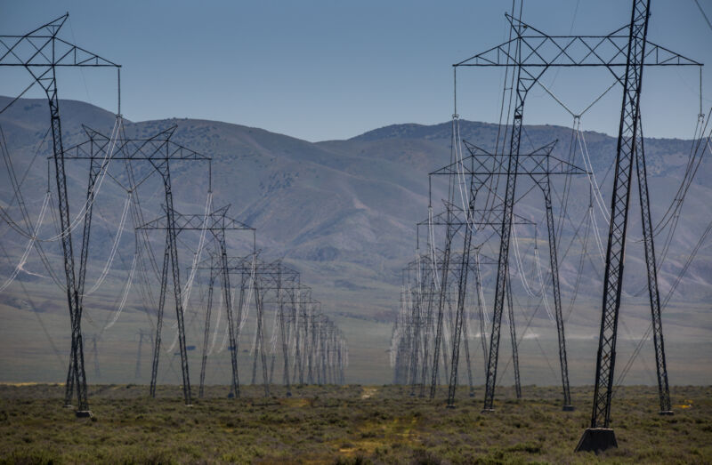 Power lines cross a green field with mountains in the background.