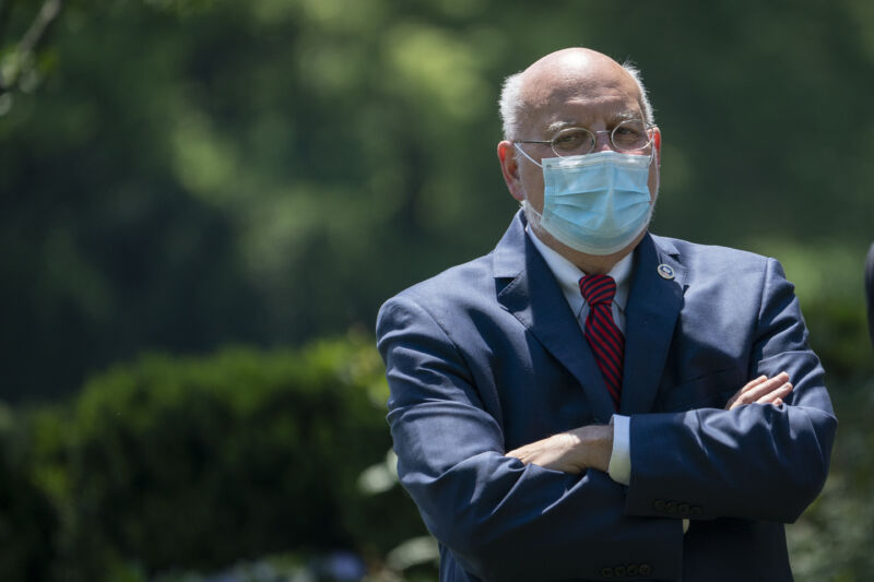 A man in a suit and face mask stands outside with his arms folded.