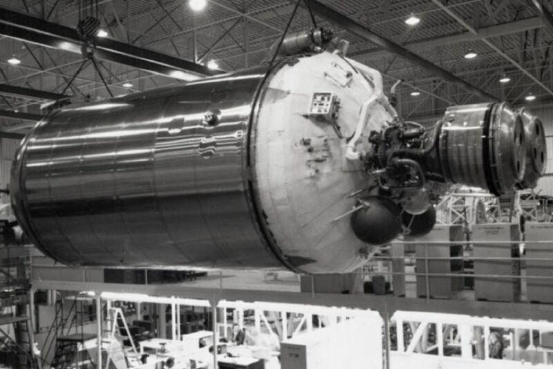 Black-and-white photo of a huge rocket component in a giant hangar.