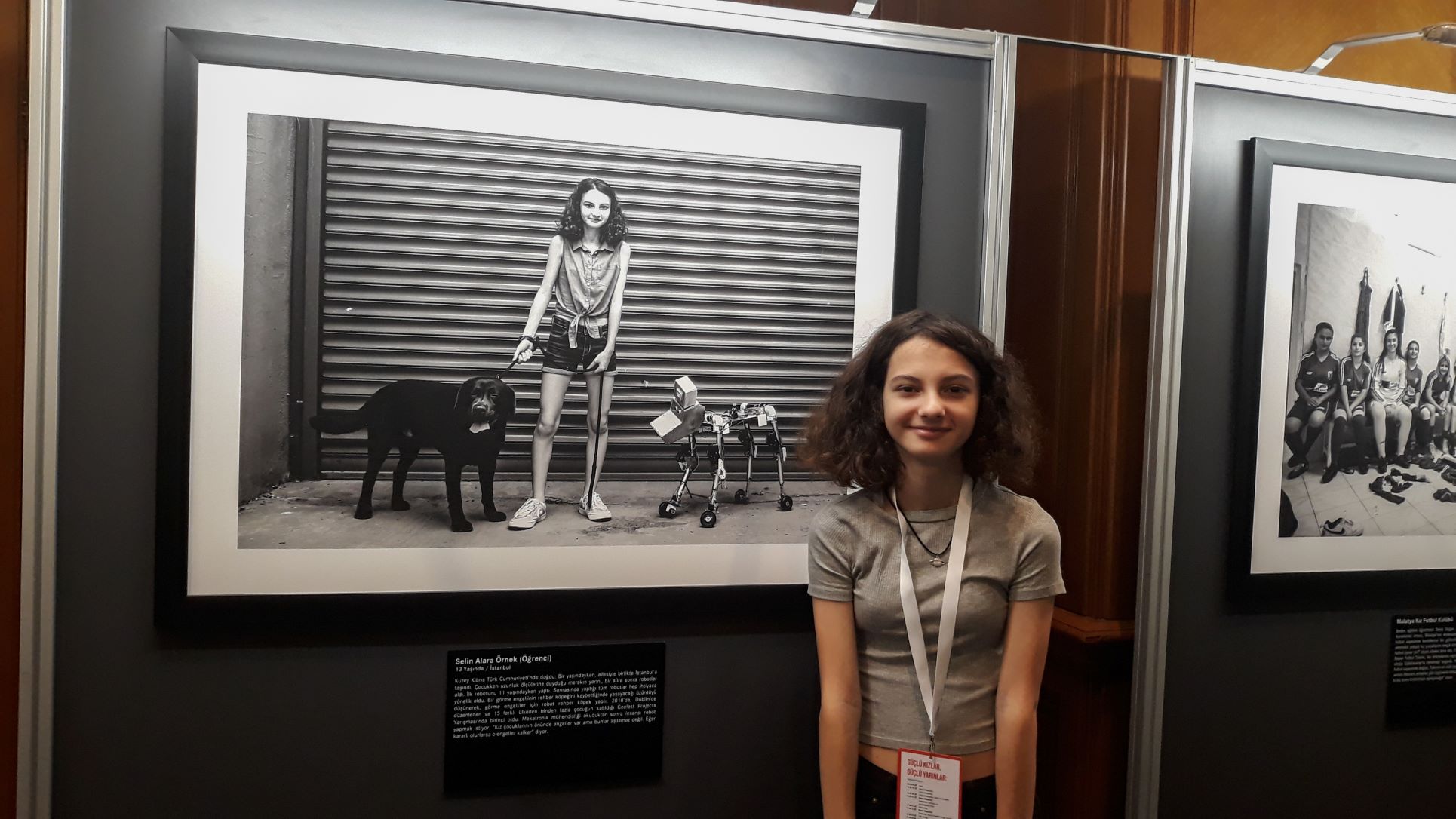 Selin stands next to an photograph of herself. In the photograph she has a dog on one side and a robot dog on the other.