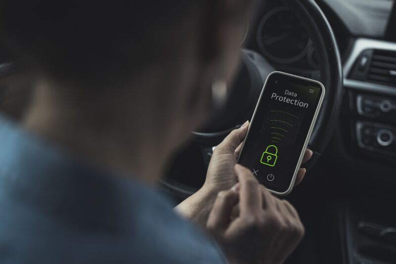 A person sits in a car holding a smartphone, the screen reads