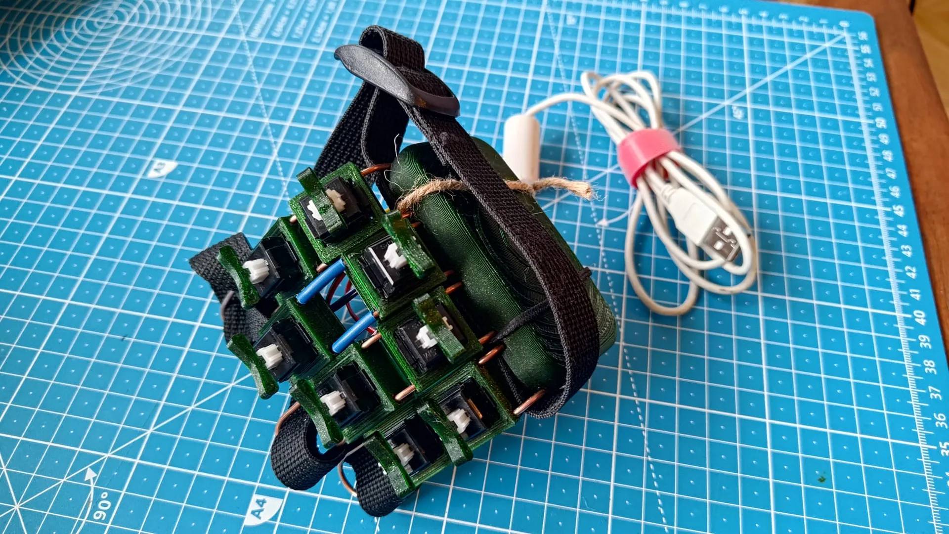 Self-built portable electronics device with several green circuit boards and black webbing on a blue cutting mat. A USB cable with a pink cable holder is ready for connection to a computer for programming or power supply.