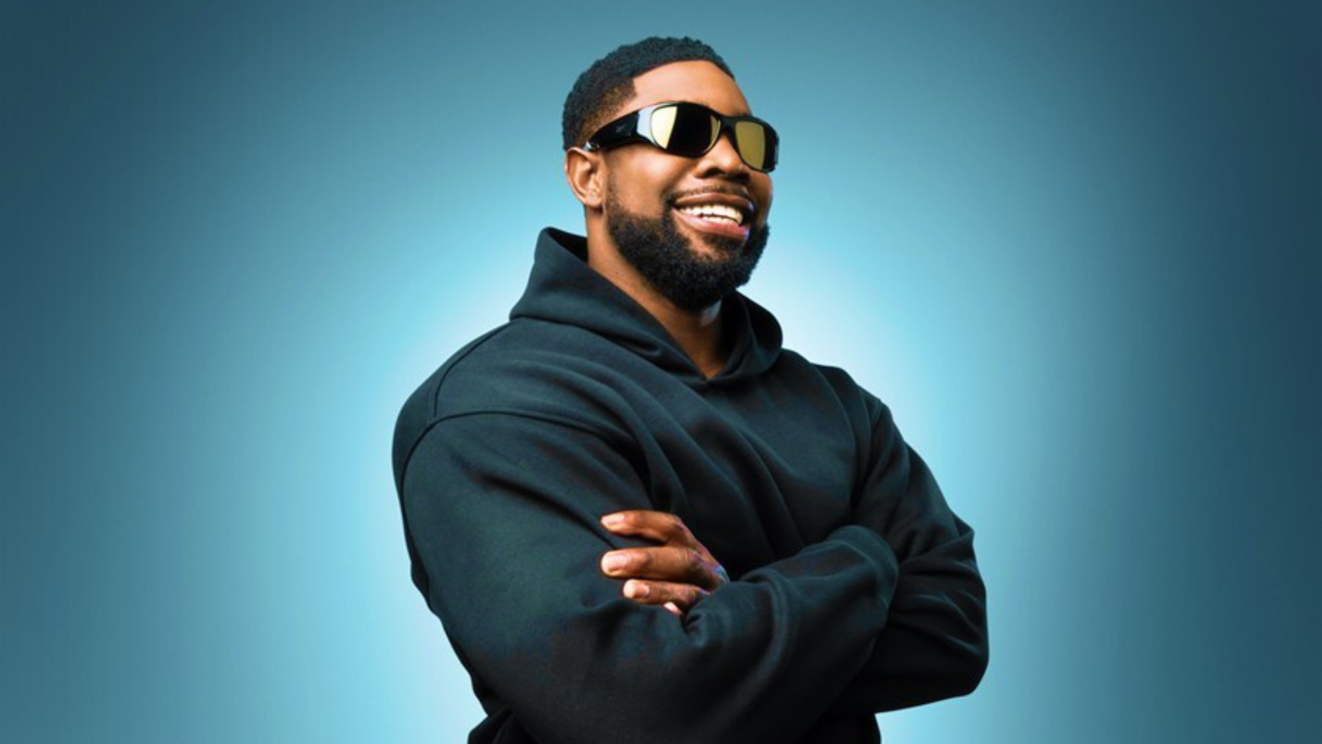 Smiling man in black hoodie and sunglasses with folded arms in front of a blue background.