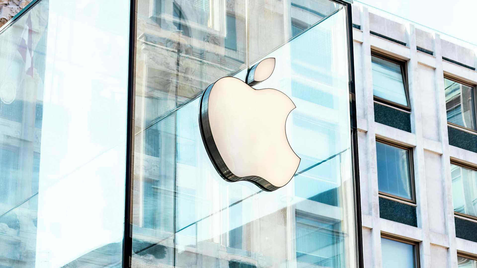 The Apple logo on the glass façade of an Apple Store reflects the surrounding architecture.