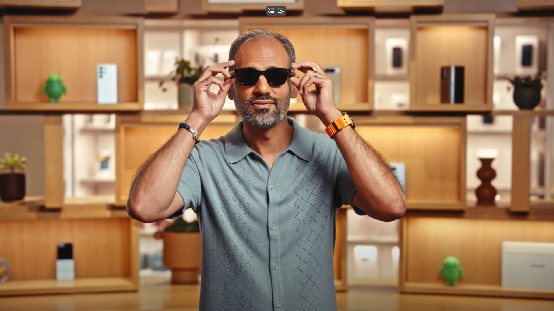 A man in a blue shirt puts on black smart glasses that look like normal sunglasses.