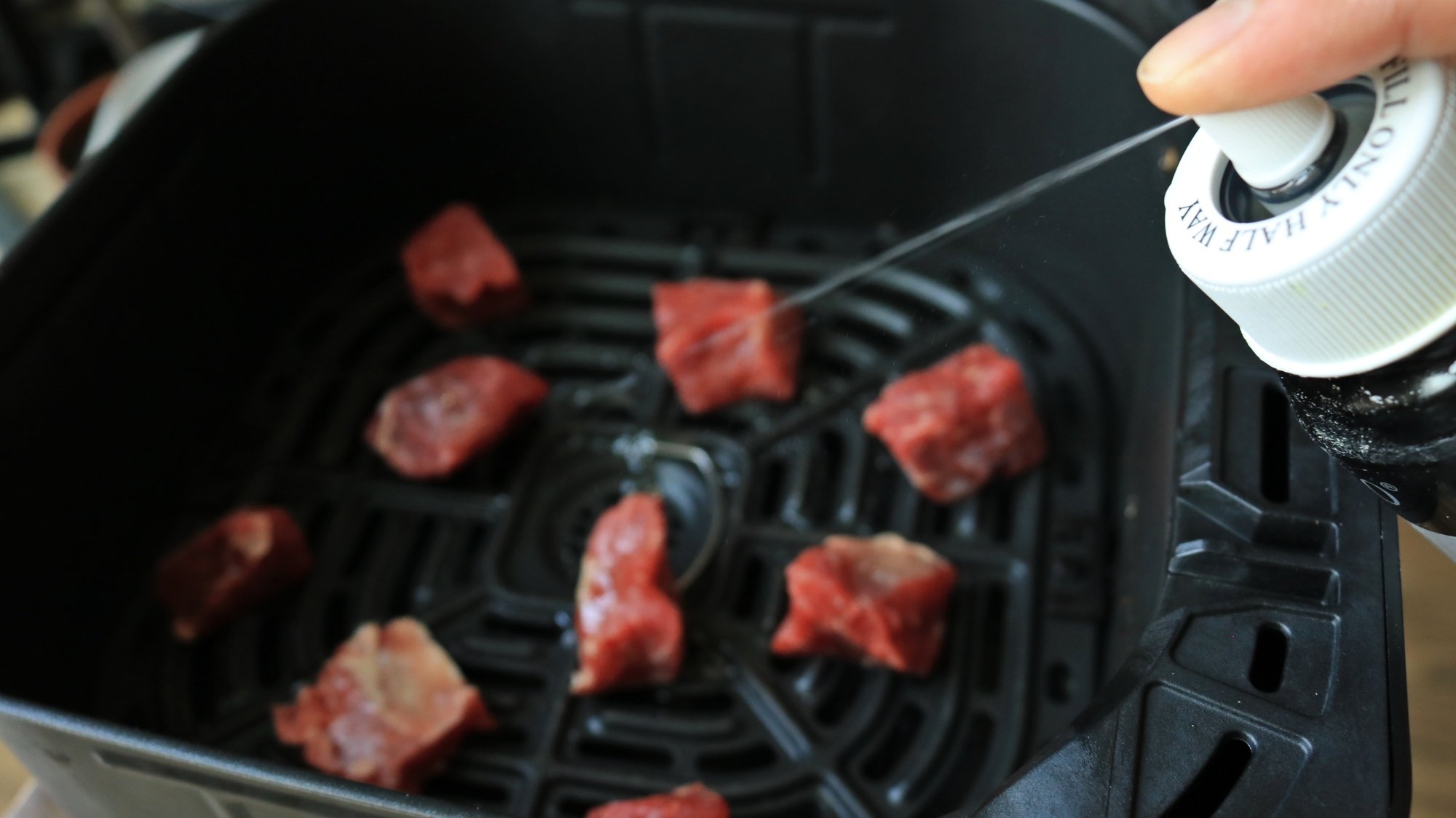 Oil canister spraying oil onto steak bites in an air fryer basket.