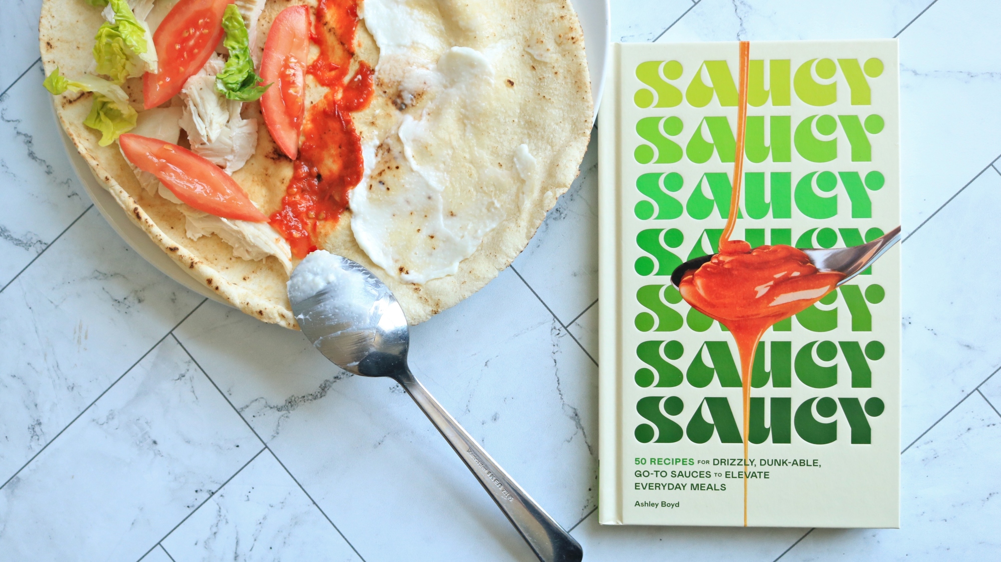 'Saucy' cookbook on a counter next to a pita with sauce and vegetables.