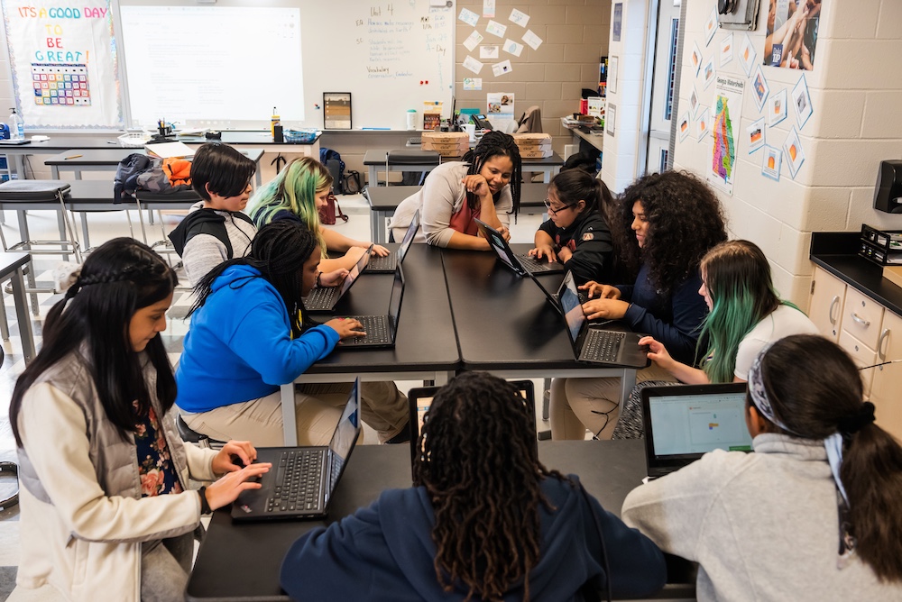 Students use their laptops in a classroom, supervised by a teacher.