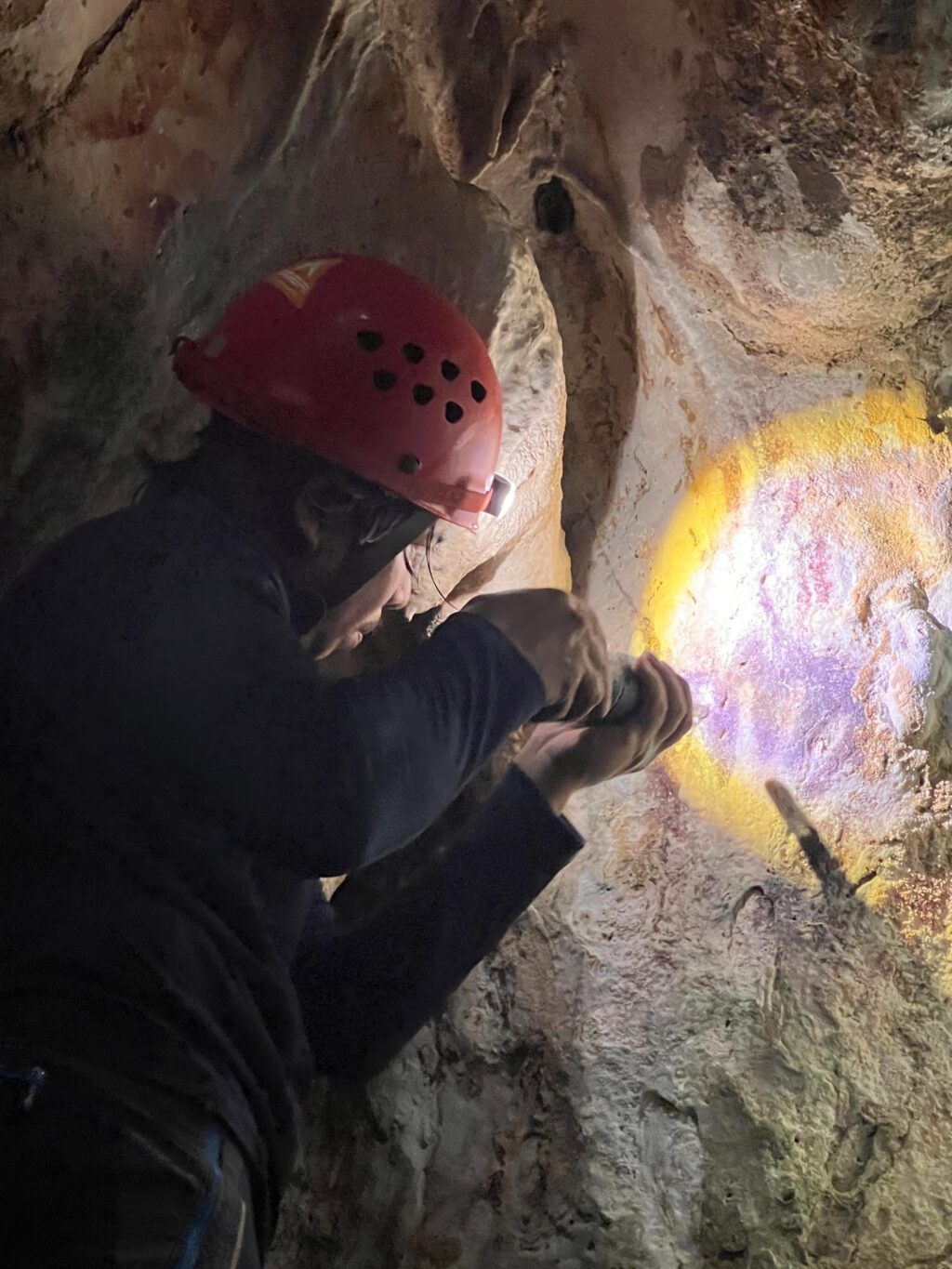 Photo of an archaeologists examining a hand stencil painted on a cave wall, using a flashlight