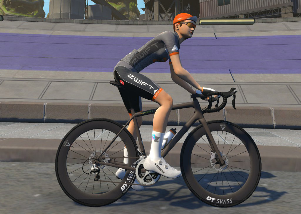 A cyclist wearing a gray Zwift jersey, black shorts, orange cap, and sunglasses rides a black bike with DT Swiss wheels on a paved road in a cityscape, with a purple ramp and concrete barriers in the background.