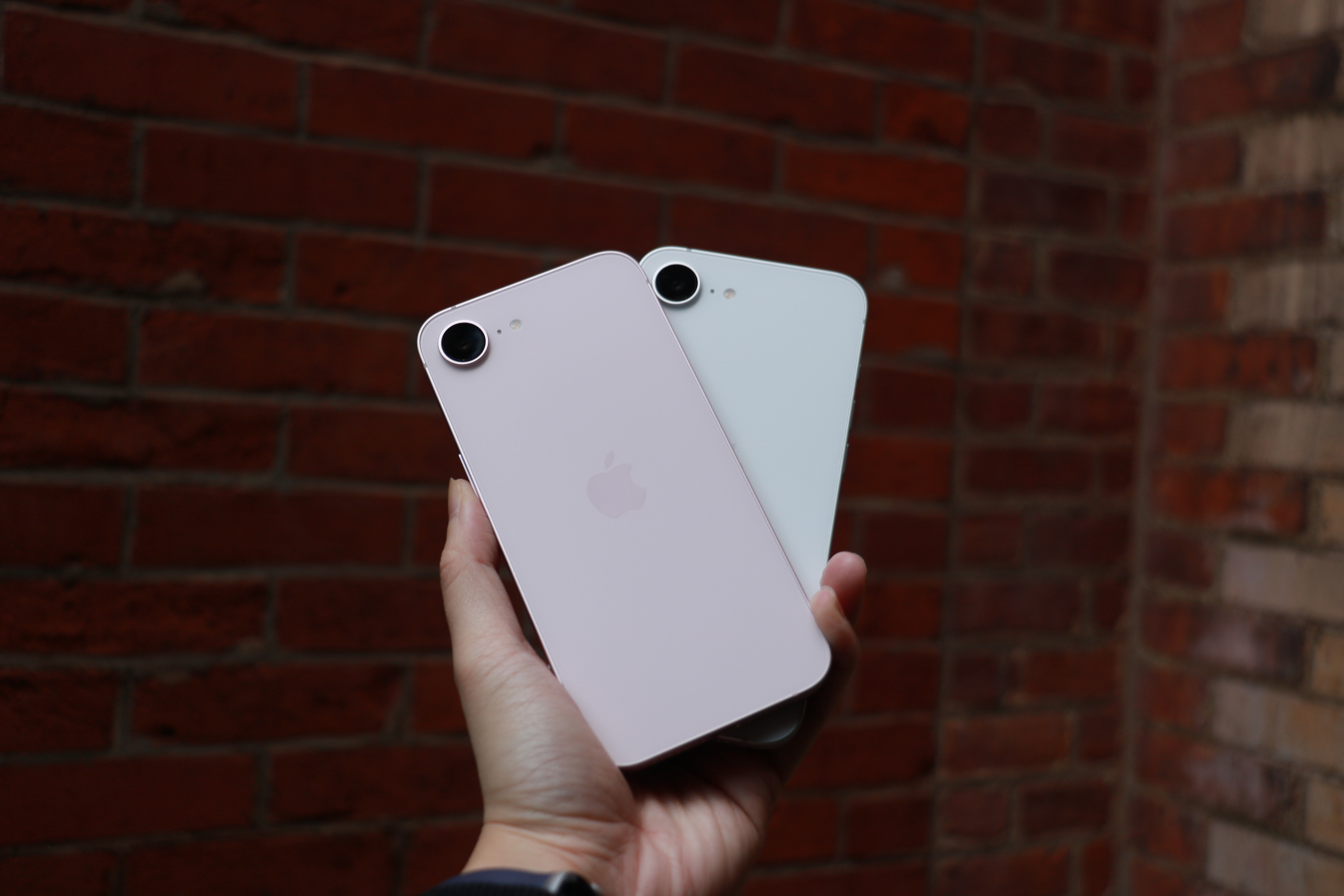 The pink iPhone 17e and the white iPhone 16e held up in one palm in front of a red brick wall.