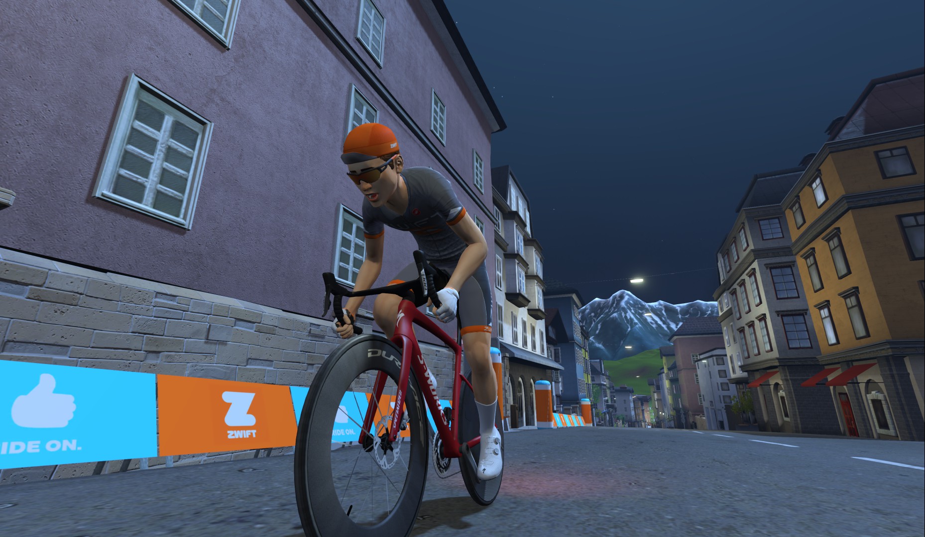 A cyclist in orange and black gear rides a red bike through a virtual city street at night, with mountains in the background and colorful banners along the roadside.